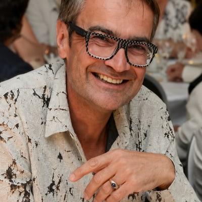 paris-die-normandie-und-das-meer-1.png Person mit Brille in weissem Hemd lächelnd bei einem formellen Essen.