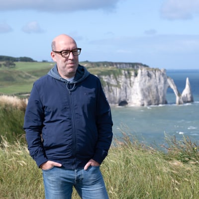 eine-sommerliche-litera-tour-2.jpg Eine Person mit Brille steht in einer grasbewachsenen Landschaft mit Meer und Felsen im Hintergrund.