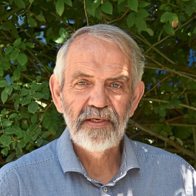 Älterer Mann mit Bart vor einer grünen Hecke.
