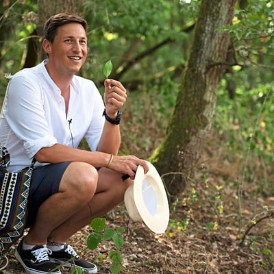 gartenwelten-an-drei-fluessen-1.jpg Mann im Wald mit Strohhut, Blatt in der Hand, lächelnd.