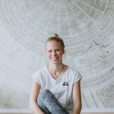 gesund-und-fit-mit-dorrit-auf-dem-rhein-1.jpg Person sitzt lächelnd auf Yogamatte vor gemustertem Hintergrund.