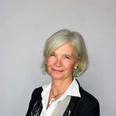 das-welterbeder-donau-1.png Porträt einer lächelnden, älteren Frau in formeller Kleidung vor grauem Hintergrund.