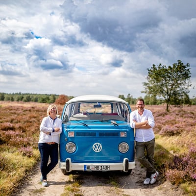 ani-und-arne-soetebier-mit-einem-der-heide-bullis.jpg Zwei Personen lehnen an einem blauen Retro-Van auf einem ländlichen Feldweg mit violetten Blumen.