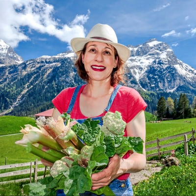 sabines-sommergaerten-1.jpg Person in rotem T-Shirt mit Hut hält Gemüse in einer Berglandschaft.