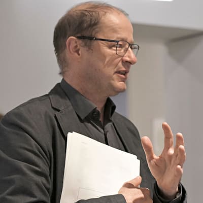 meisterwerke-von-paris-zum-meer-1.jpg Person in schwarzem Anzug hält Papiere und gestikuliert während des Sprechens.