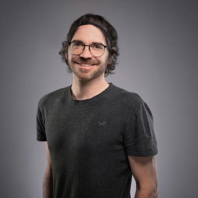 fotoexperte-stefan-tschumi-nikon-school.jpg Mann mit Brille und lockigem Haar lächelt vor grauem Hintergrund.