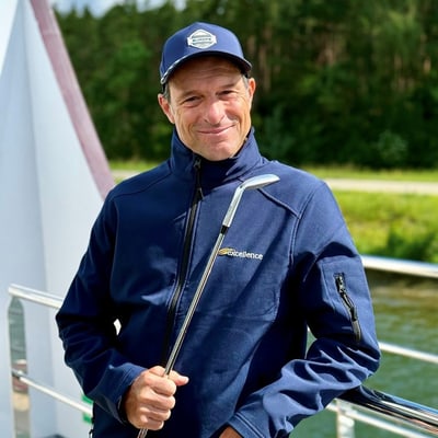 die-gruensten-seiten-der-donau-1.jpg Person in blauer Jacke und Mütze steht mit einem Golfschläger auf einem Boot.