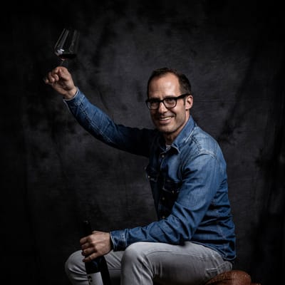 Mann in Jeansjacke lächelt, hält Weinglas und Flasche, sitzt auf Stuhl.