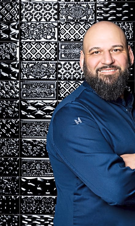 Mann in blauem Hemd vor einer Wand mit dekorativen schwarzen Fliesen.