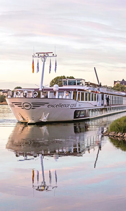 Flusskreuzfahrtschiff Excellence Coral mit weissem Rumpf und lila Akzenten gleitet bei Sonnenuntergang an einer Stadt mit Kirchturm vorbei.