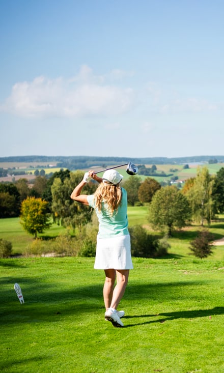 Frau in hellgrünem Shirt und weissem Rock führt einen Golfschlag auf einem weitläufigen Golfplatz aus.