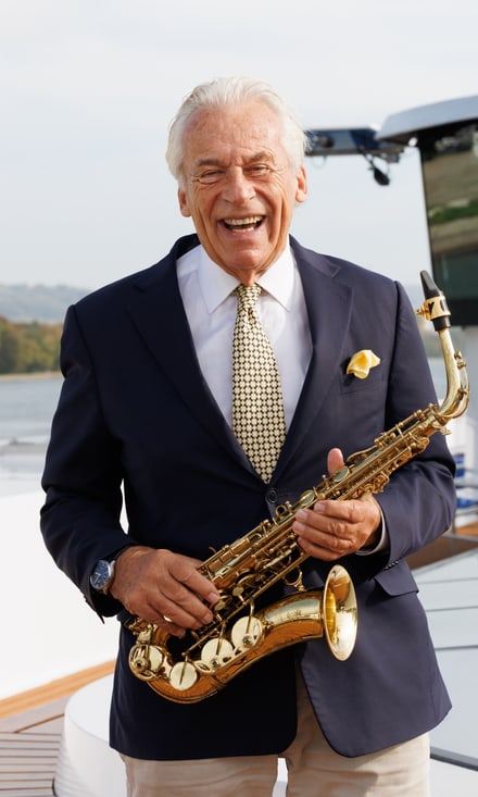 Pepe Lienhard im blauen Anzug hält ein goldenes Saxophon und lächelt auf dem Sonnendeck eines Flussschiffs.