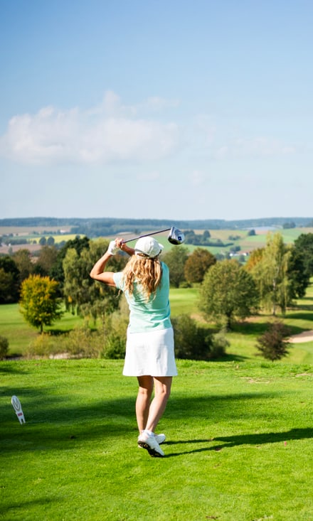 Frau in hellgrünem Shirt und weissem Rock führt einen Golfschlag auf einem weitläufigen Golfplatz aus.