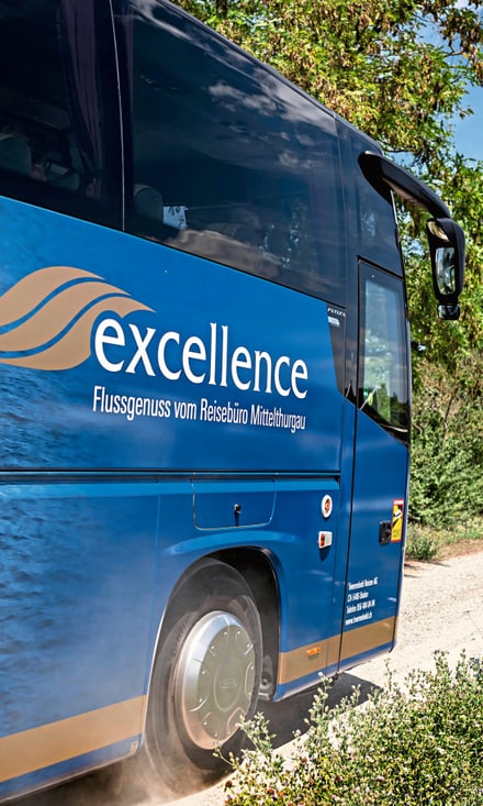 Blauer Excellence-Bus am Flussufer, im Hintergrund das Flusskreuzfahrtschiff Excellence Baroness am Steg.
