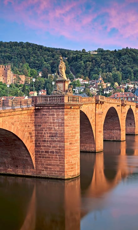 twerenbold_78079.jpg Historische Steinbrücke über einen Fluss vor einer malerischen Altstadt und Hügeln bei Sonnenuntergang.