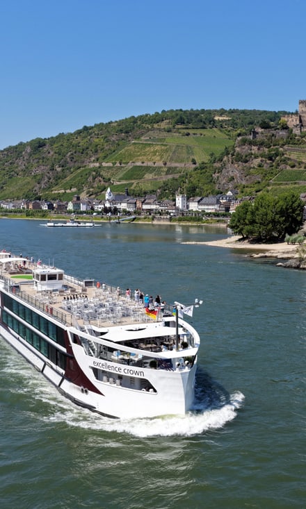 Das Flussschiff Excellence Crown fährt auf dem Rhein, Passagiere auf dem Sonnendeck geniessen den Blick auf Weinberge und historische Gebäude.