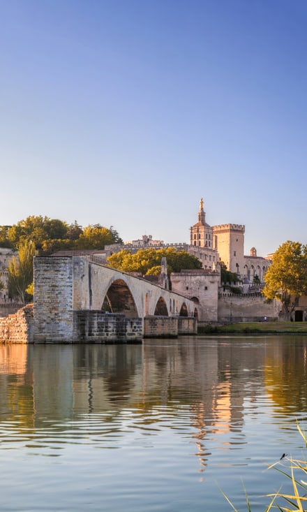 Steinerne Bogenbrücke Pont d'Avignon spiegelt sich im Rhône, dahinter der historische Papstpalast unter blauem Himmel.