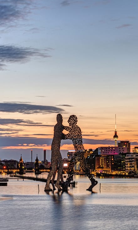 Zwei punktierte Silhouetten-Skulpturen spiegeln sich im Abendlicht in der Spree, dahinter der Berliner Fernsehturm.