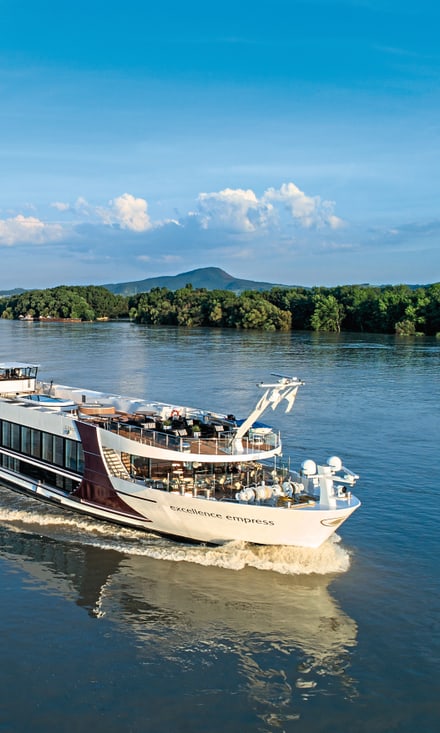Das Flusskreuzfahrtschiff Excellence Empress fährt auf einem ruhigen Fluss, Gäste stehen und sitzen auf dem Sonnendeck.