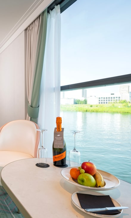 Helle Kabinen-Suite mit Doppelbett, Sofa, Sessel und grossen Fenstern mit Blick auf grünes Flussufer.