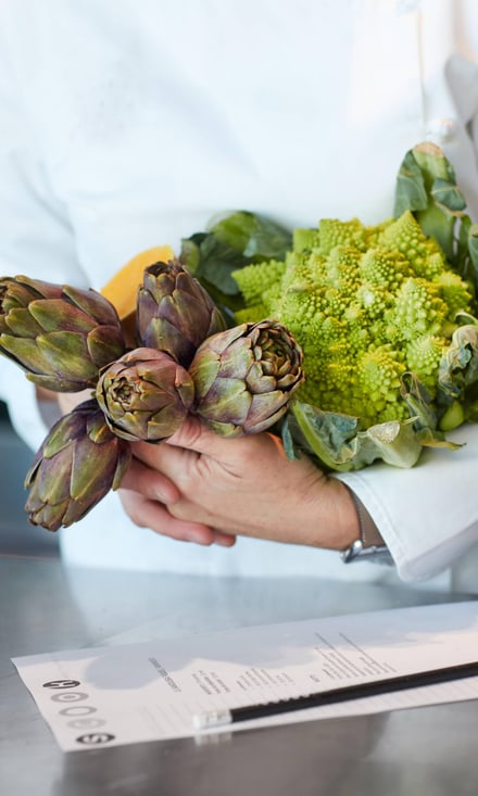 Koch hält Artischocken und Romanesco im Arm neben Rezeptblatt.