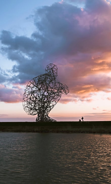 Grosse Metallskulptur bei Sonnenuntergang neben Wasser.