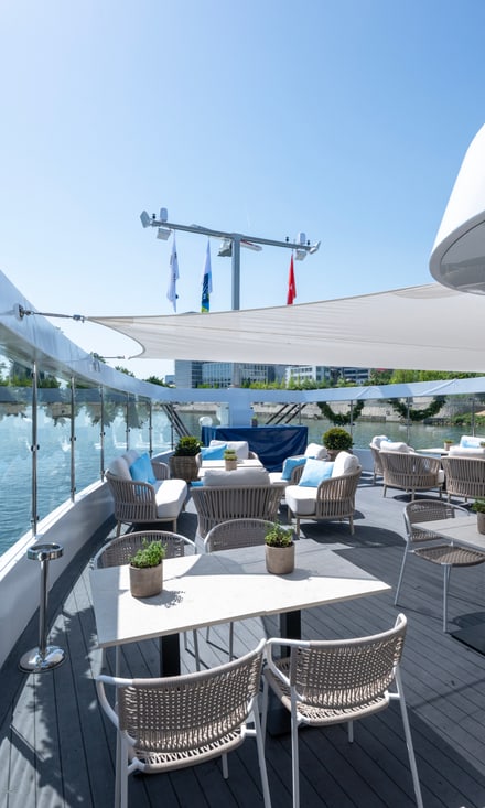 Leeres Sonnendeck der Excellence Crown mit weissen Tischen, Korbstühlen und Blick auf einen Fluss mit Stadtkulisse.