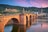 Historische Steinbrücke über einen Fluss vor einer malerischen Altstadt und Hügeln bei Sonnenuntergang.