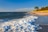 Wellen am Sandstrand mit grüner Küste im Hintergrund.
