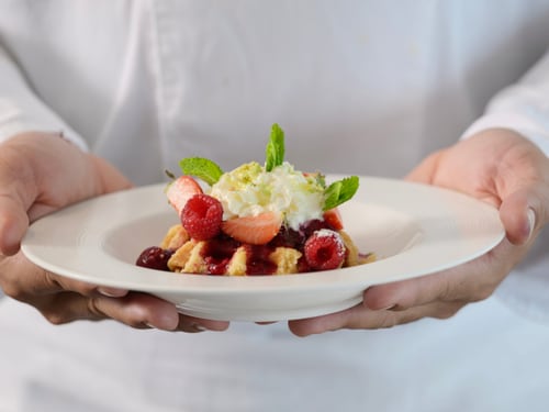 Person in weissem Hemd hält Teller mit Dessert, garniert mit Früchten und Minzblättern.