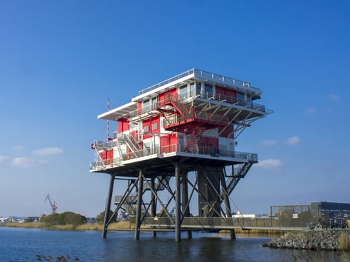 Mehrstöckiges Gebäude auf Stahlstützen über dem Wasser im Hafen von Amsterdam, mit Geländern und Treppen.