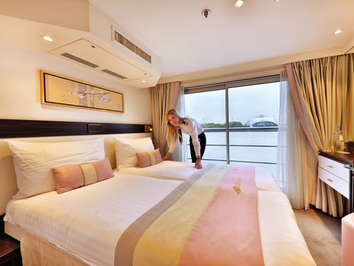 Crewmitglied richtet Bett in Kabine der Excellence Pearl, Blick durch Panoramafenster auf Fluss und Ufer.