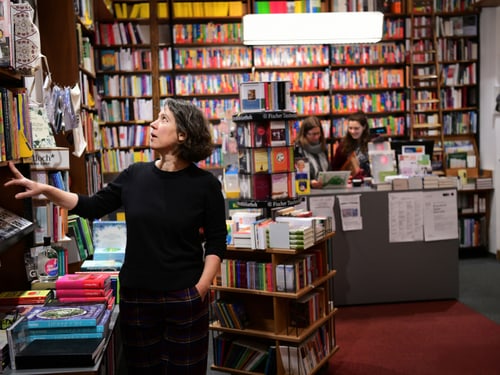 Frau wählt Buch in einem Buchladen, im Hintergrund zwei Frauen an der Kasse.
