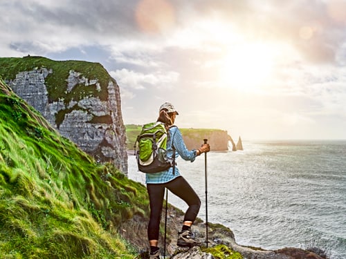 Frau mit Rucksack und Trekkingstöcken steht auf felsigem Vorsprung an der Küste der Normandie mit Meeresblick.