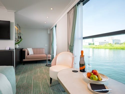 Helle Kabinen-Suite mit Doppelbett, Sofa, Sessel und grossen Fenstern mit Blick auf grünes Flussufer.