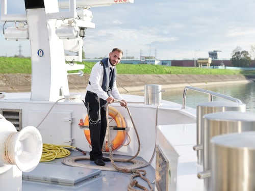 Mann in Schiffsuniform hält Seile auf dem weissen Deck eines Fluskreuzfahrtschiffs bei sonnigem Wetter.
