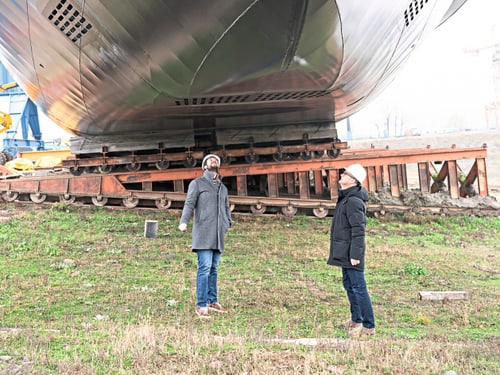 Zwei Arbeiter mit Schutzhelmen betrachten ein grosses zylindrisches Bauteil auf rotem Transportgestell auf dem Werftgelände.