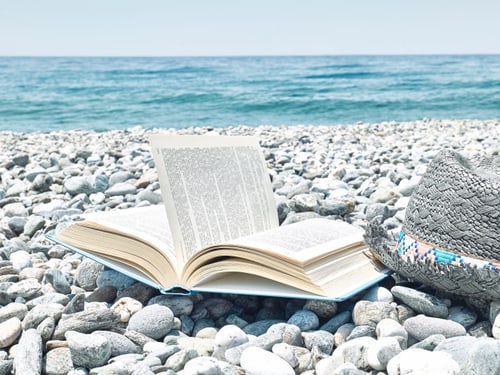 Offenes Buch und Hut auf kieseligem Strand am Meer.