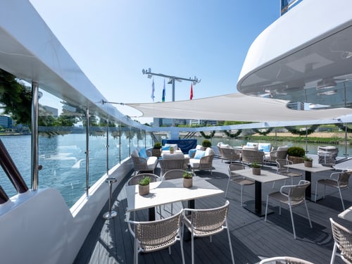 Leeres Sonnendeck der Excellence Crown mit weissen Tischen, Korbstühlen und Blick auf einen Fluss mit Stadtkulisse.