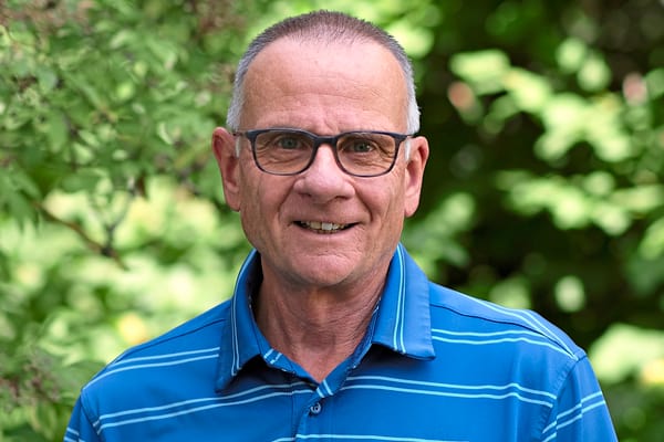 Älterer Mann mit Brille in blauem Poloshirt vor grünem Hintergrund.