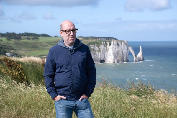 Eine Person mit Brille steht in einer grasbewachsenen Landschaft mit Meer und Felsen im Hintergrund.