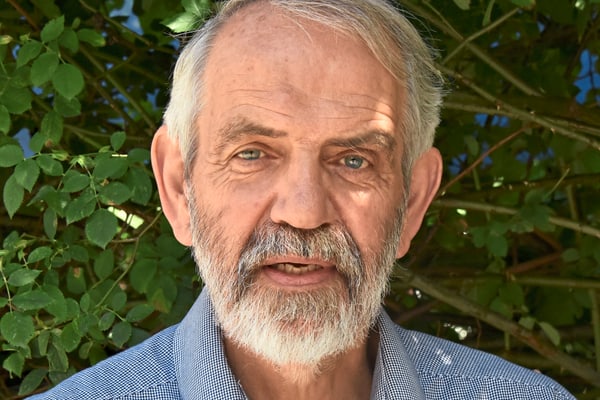 Älterer Mann mit Bart vor einer grünen Hecke.