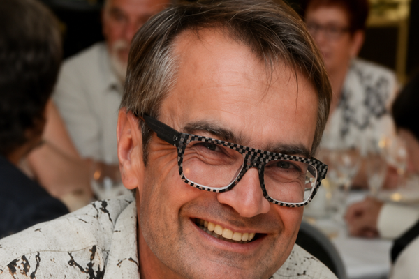 paris-die-normandie-und-das-meer-1.png Person mit Brille in weissem Hemd lächelnd bei einem formellen Essen.