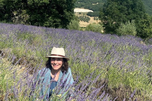 lavendelbluete-in-der-provence-1.jpg Eine Person mit Hut sitzt in einem Lavendelfeld mit Bäumen und Hügeln im Hintergrund.