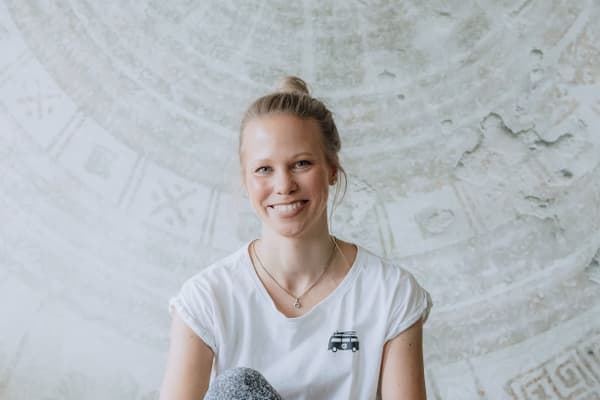 Person sitzt lächelnd auf Yogamatte vor gemustertem Hintergrund.