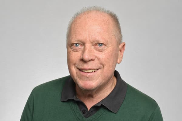 Älterer Mann in grünem Pullover vor grauem Hintergrund.