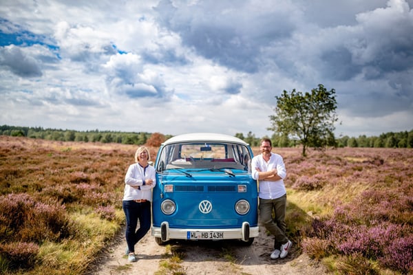 ani-und-arne-soetebier-mit-einem-der-heide-bullis.jpg Zwei Personen lehnen an einem blauen Retro-Van auf einem ländlichen Feldweg mit violetten Blumen.