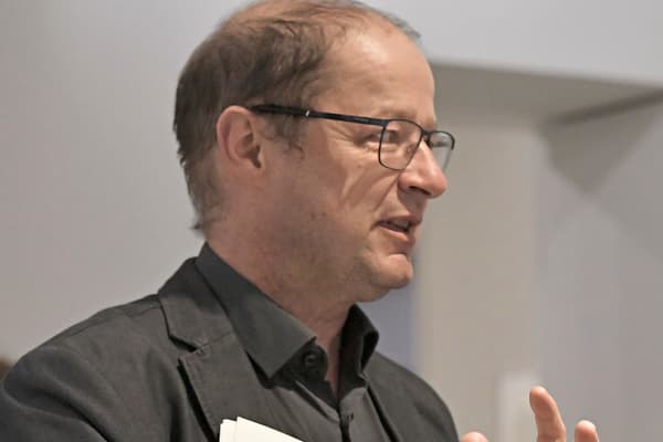 Person in schwarzem Anzug hält Papiere und gestikuliert während des Sprechens.