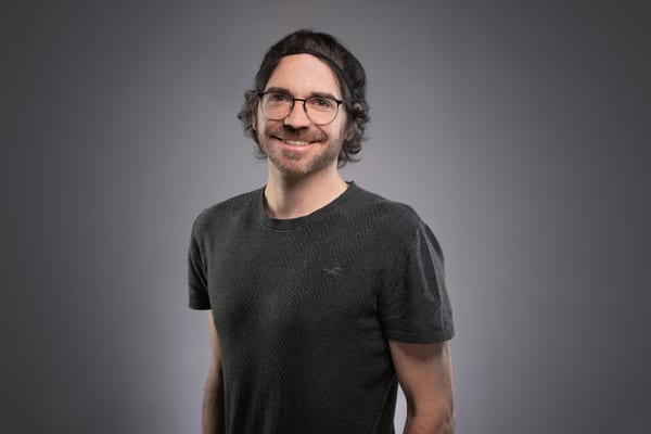 Mann mit Brille und lockigem Haar lächelt vor grauem Hintergrund.