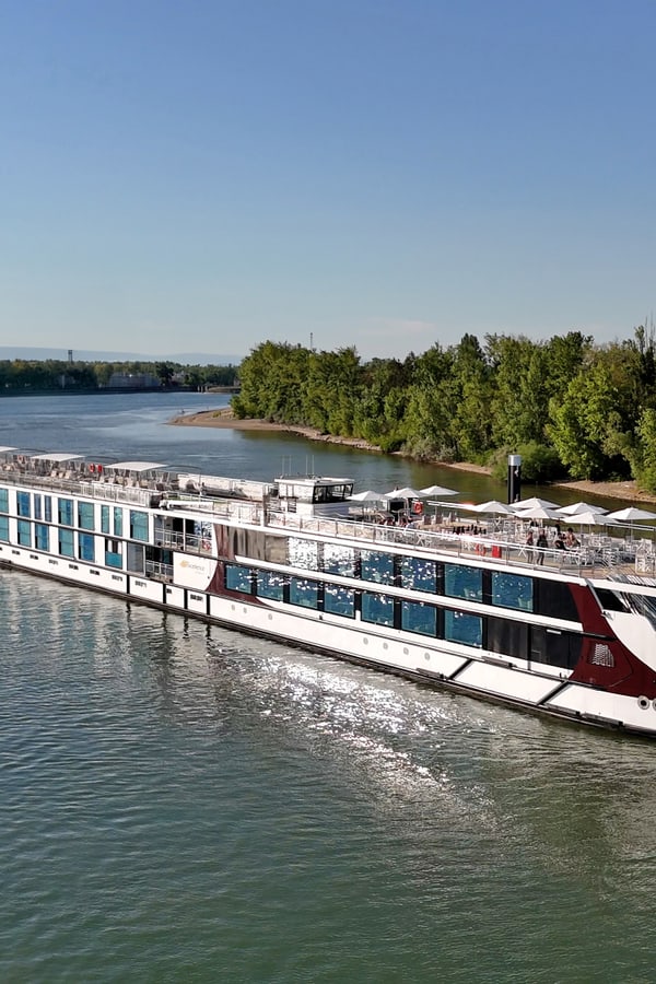 Das Flusskreuzfahrtschiff Excellence Crown liegt an einem grünen Flussufer, Gäste geniessen das Sonnendeck.